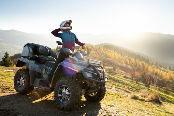 Young happy woman in protective helmet enjoying extreme ride on atv quad motorbike in autumn mountains at sunset.
