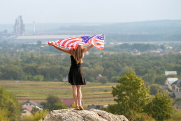 Young happy american woman with long hair raising up waving on wind USA national flag in her hands relaxing outdoors enjoying warm summer day.