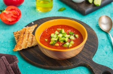 Tomato gazpacho, traditional Spanish cold soup or appetizer in a yellow clay bowl on a wooden board against a turquoise concrete background. Cold soup recipes.