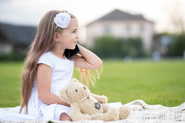 Pretty child girl sitting in summer park on green grass with her teddy bear toy talking on mobile...