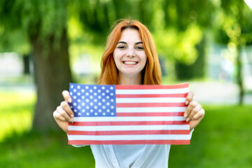 Portrait of happy young red haired woman holding USA national flag in her hands standing outdoors in summer park. Positive girl celebrating United States independence day.