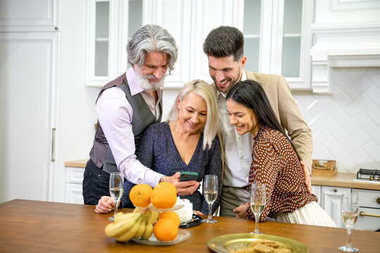Positive grandparents and smiling young couple in formal wear looking at pictures and videos on smartphone during birthday celebration with family at home in kitchen with champagne on table