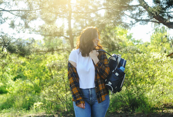 Smiling happy woman in yellow checkered shirt with a traveling bag spends time in the forest. Summer time holidays concept. 