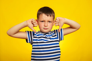 Obraz premium A young boy in striped t-shirt showing thumbs down gesture with two hands on yellow background. Displeased child giving thumbs down hand gesture