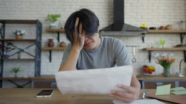 Young tired man scratching head analyzing business graph in home office. Portrait of troubled Asian manager thinking planning strategy idea indoors in kitchen. Entrepreneurship concept