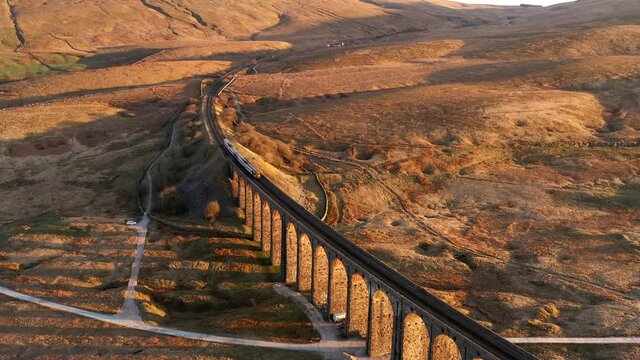 Aerial 4K drone footage showing train crossing over Ribblehead Viaduct in the Yorkshire Dales. Taken in the morning golden hour with stunning warm colours from the rising sun.
