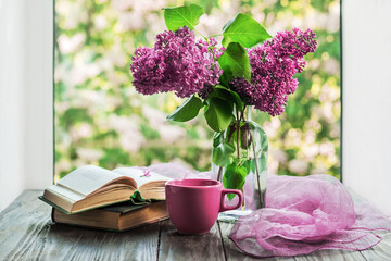 Morning coffee. A cup of coffee or tea, a bouquet of purple lilacs and an open book on a wooden table with a view out the window. Still life concept, blurred background.