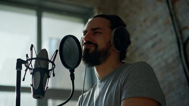 A young bearded man with headphones sings into a microphone on a stand with a pop filter, singer records a song at a recording studio.