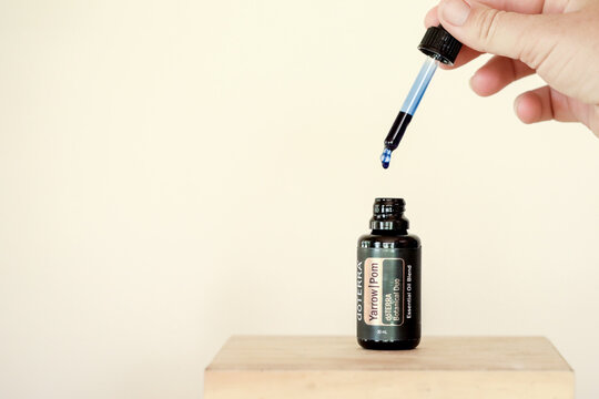 Illustrative editorial image of doTERRA Yarrow Pom Essential Oil on plain white background with hand holding dropper full of blue liquid