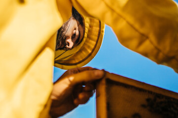 Close portrait shot from ground of a young beekeper with a beard in a yellow protecting suit...
