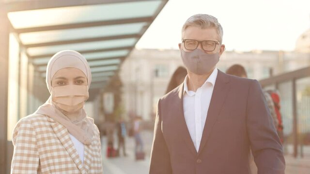 Close up portrait of mixed-race people in protection masks standing in city on street and looking at camera. Business trip. Businessman and business lady outdoors at bus station Covid pandemic concept