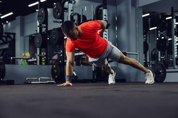 The whole body and core balance of a shaped man. He is in a plank position with one hand on the floor and one on his back. He wears sportswear and an armband. Sport, love, motivated