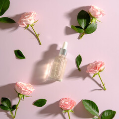 Transparent glass dropper bottle with serum and rose flowers on pink background. Flat lay, top view.