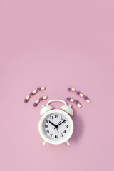 White alarm clock surrounded by multi-colored pills on pink background. Time to take vitamins concept. Vertical banner. Copy space.