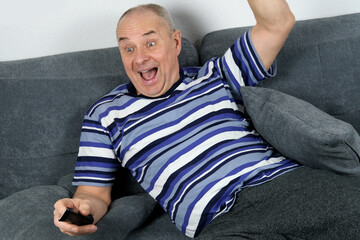 Fototapeta premium old bald man sitting on a sofa and watching tv, emotionally screams, remote control in hand, concept of a cozy home evening, family life