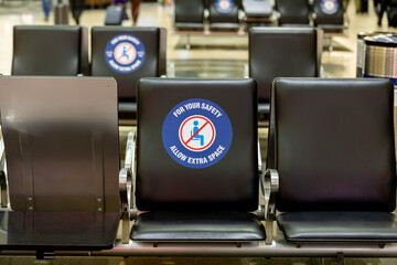 Black seat in the airport terminal with large poster read "For your safety allow extra space". An sign to prohibit seat occupation during COVID lockdown
