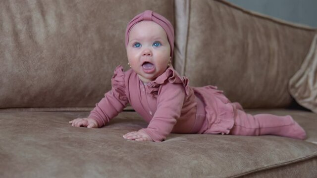 A baby girl with blond hair and blue eyes in pink clothes lies on her tummy and actively moves her arms and legs. Baby on a light beige sofa