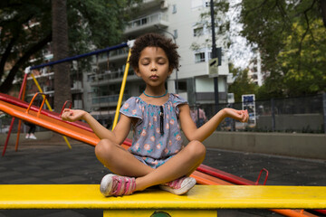 Mixed race girl relaxing and concentrated with eyes closed doing yoga and meditation gesture with fingers in lotus pose at playground outdoors.