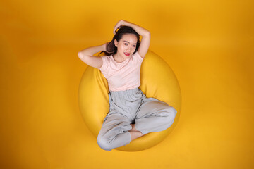 Beautiful young south east Asian woman sits on a yellow beanbag seat orange yellow color background pose fashion style elegant beauty mood expression rest relax think emotion top view