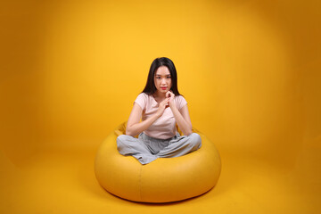 Beautiful young south east Asian woman sits on a yellow beanbag seat orange yellow color background pose fashion style elegant beauty mood expression rest relax exercise stretch yoga