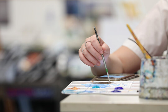 A student's hand isolated holding a paint brush mixing paints at a school desk in the visual art room. A mobile phone is on the desk either as research or distraction tool.