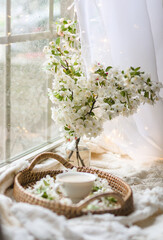 Fototapeta na wymiar Fresh Spring blossom with a cup of coffee near window with raindrops where sheer curtain and string lights are hanging