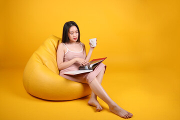 Beautiful young south east Asian woman sit on a yellow orange beanbag seat color background relax read study think book magazine hold white coffee cup