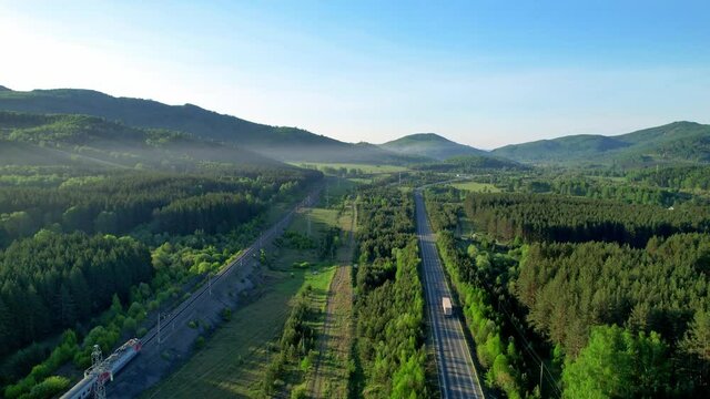 Simultaneous movement of trains and trucks parallel to each other in a picturesque location. Railway tracks and a highway in the middle of the forest and mountains. View of the road from the height.
