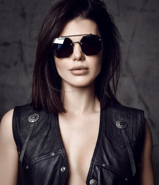 Portrait of powerful brutal sexy pretty brunette woman in black leather vest on naked body and sunglasses over grey concrete wall background. Beautiful women and sexual outfit concept