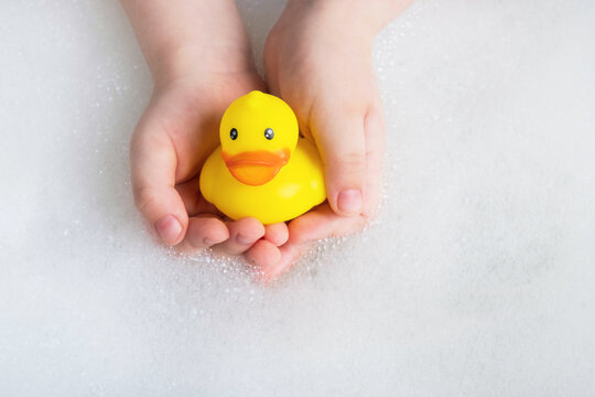 Little Child Holding Yellow Rubber Duck In Bubble Bath. Baby Hygiene, Accessories For Bathing, Toys. Top View, Close Up