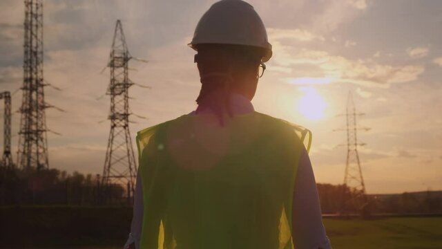 Woman engineer walking across  field with high voltage electrical pylons at sunset. Ensuring uninterrupted supply and distribution of electricity to consumers.