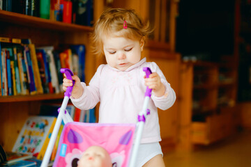 Cute adorable baby girl making first steps with doll carriage. Beautiful toddler child pushing stroller with toy at home. Happy daughter learning walking and standing