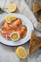 Gravlax Graved Lachs, ein roh gebeizter Fisch mit Dill auf Emaille Teller mit Hovmästarsås Senf Soße, Toast, Zwiebel Ringe, Zitrone und Leinen Tuch auf hell Marmor Hintergrund