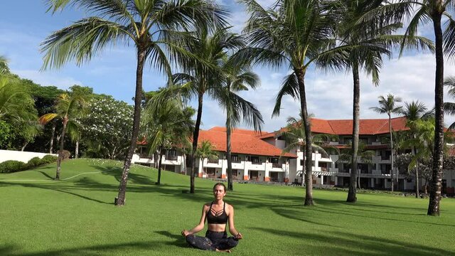 Sporty girl sitting in lotus pose doing yoga namaste munda hands pose on green grass in park with palm trees. Yoga woman outdoor. Healthy lifestyle concept