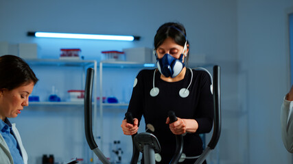 Team of doctor researchers monitoring endurance of woman performance sports. Athlete wearing mask running cross trainer while scientist talks with her measuring vo2, health status, physical enducance