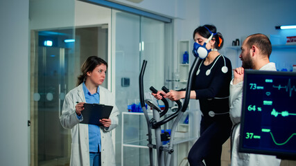 Sportswoman running with mask on cross trainer with electrodes attached to body. Physician using tablet computer and controls EKG data showing on laboratory monitors, discussing with patient