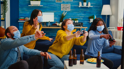 Excited multiethnic friends trying to win video games enjoying new normal party during global pandemic wearing face mask, keeping distancing sitting on couch in living room supporting women
