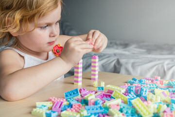 Small preschooler girl playing with colorful toy building blocks, sitting at the table.