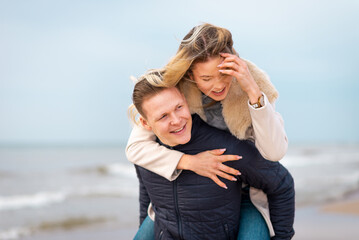 Happy man giving piggyback ride to his woman and laughing at beach. Smiling guy in love carrying on...