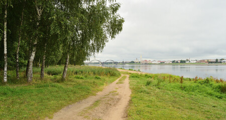 The bank of the river, a birch grove, a dirt road leads into the distance, where on the opposite bank against the background of the sky you can see the old city. Cloudy summer day.