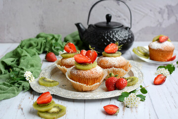 Muffins decorated with strawberries and kiwi, black teapot and green napkin on a wooden background