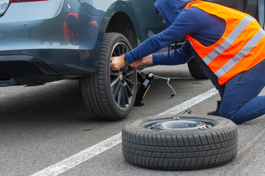 A man in a orange safety vest changes a flat tire on a road. Closeup mans hands to the wheel of a broken car. Replacement of a wheel using skrewdriver, Prague, March, 2020