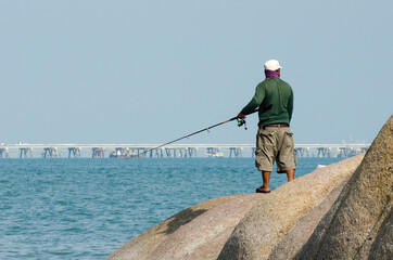 An unknown man standing fishing on a rock in the middle of the sea, industrial estate background.