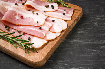 Board with raw bacon with spices, and rosemary on a black background