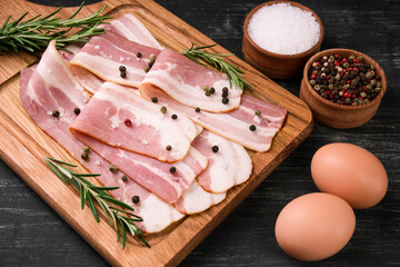 Board with raw bacon with spices, and rosemary on a black background