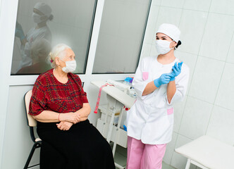 a nurse in a medical mask puts on rubber gloves and prepares for a medical examination of a patient - an elderly gray-haired woman