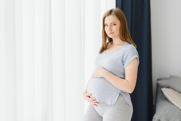 Beautiful pregnant woman is touching her belly and smiling while standing near the window at home