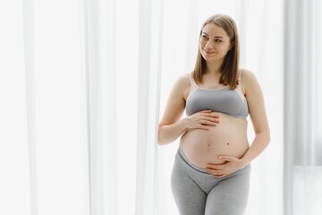 Young beautiful pregnant woman standing at home near the window.