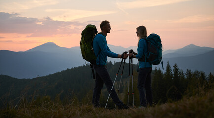 Full length of smiling man and woman travelers hiking together in mountains. Silhouette of happy young couple with backpacks standing on grassy hill with sunset and grassy hills on background.