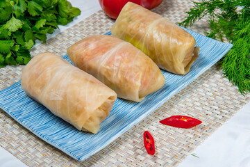 Three raw stuffed cabbage rolls on a blue rectangular plate with some tomatoes dill and parsley aside